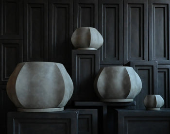 Four 101 Copenhagen Arket Plant Pot, Petit vases by 101 Copenhagen are displayed on dark geometric pedestals before a paneled wall, creating a minimalist and dramatic arrangement.