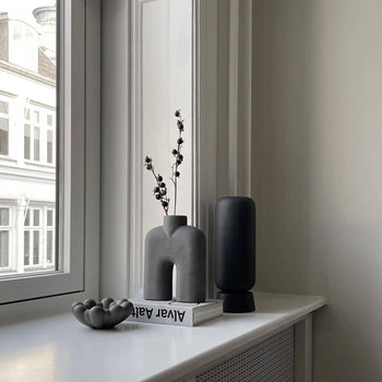 A minimalist windowsill displays a sculptural gray vase with dried branches, the 101 Copenhagen Bloom Tray, Mini, a stack of books, an organic-shaped sculpture, and a tall black vase, with buildings visible through the window.