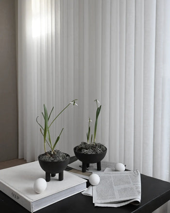 The 101 Copenhagen Duck Bowl, Big by 101 Copenhagen sits on a dark table beside white ping pong balls and open newspapers. Sheer white curtains in the background diffuse soft light, enhancing the Scandinavian-inspired scene.