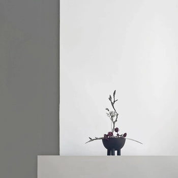 A minimalist arrangement of dark red flowers and green stems in a small black vase sits on a white surface against a gray and white background, paired with the 101 Copenhagen Duck Bowl, Mini—ideal for elevating your home decor.