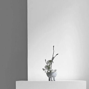 A minimalist scene features a small gray vase with delicate branches next to the 101 Copenhagen Duck Bowl, Mini, by 101 Copenhagen, elegantly displayed on a white surface against a light gray and white backdrop with clean lines.