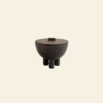 The 101 Copenhagen Duck Jar, Medio by 101 Copenhagen is a dark, round lidded jar with three cylindrical legs and a refined hand-glazed finish, offering unique decorative storage against a plain, light beige background.