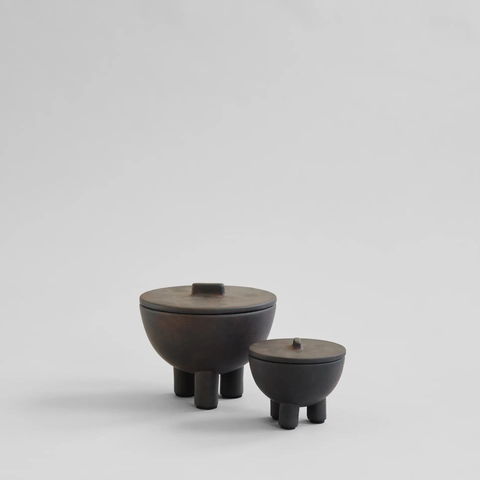 Two dark, round 101 Copenhagen Duck Jar, Medio containers by 101 Copenhagen, each with lids and three short legs, rest on a neutral light gray background. Their hand-glazed minimalist design offers stylish decorative storage.