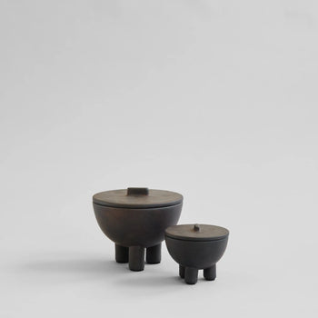 Two dark, round 101 Copenhagen Duck Jar, Medio containers by 101 Copenhagen, each with lids and three short legs, rest on a neutral light gray background. Their hand-glazed minimalist design offers stylish decorative storage.