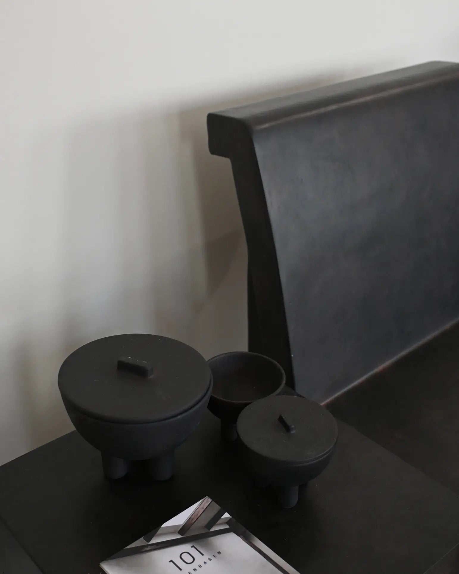 A minimalist black bench sits by a dark table adorned with three 101 Copenhagen Duck Jars, Medio—matte black, hand-glazed pieces in varying sizes for stylish storage—plus a partially visible booklet in the foreground.