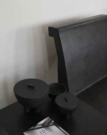 A minimalist black bench sits by a dark table adorned with three 101 Copenhagen Duck Jars, Medio—matte black, hand-glazed pieces in varying sizes for stylish storage—plus a partially visible booklet in the foreground.