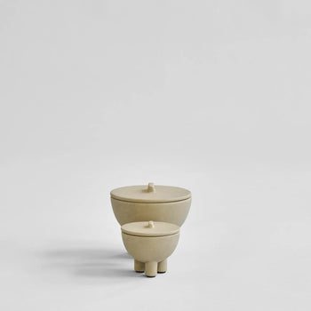 Two minimalist 101 Copenhagen Duck Jar, Medio ceramics, each beige with lids and three short legs, sit on a plain light grey background. These hand-glazed jars offer decorative storage, with the larger positioned behind and left of the smaller.