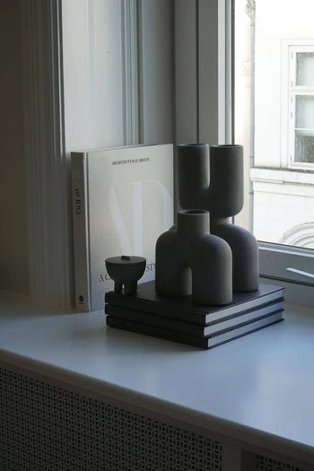 A minimalist windowsill features stacked books, sculptural gray vases, the 101 Copenhagen Duck Jar, Mini by 101 Copenhagen, a small bowl, and a large book titled Architectural Digest propped upright in natural light.