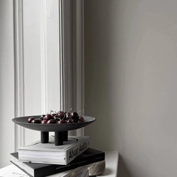 The 101 Copenhagen Duck Plate, Mini by 101 Copenhagen sits atop a stack of books near a bright window, filled with cherries and accompanied by a spool of twine—a minimalistic, softly lit accent for charming home decor.