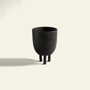 The 101 Copenhagen Duck Pot by 101 Copenhagen is a minimalist black, cup-shaped vessel with three cylindrical legs, casting a shadow on a plain light background and exuding Scandinavian design with subtle whimsical charm.