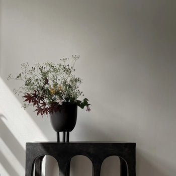 The 101 Copenhagen Duck Pot by 101 Copenhagen, filled with delicate white and burgundy flowers, sits on a dark arched table, adding whimsical charm against a light gray wall where sunlight casts elegant shadows.