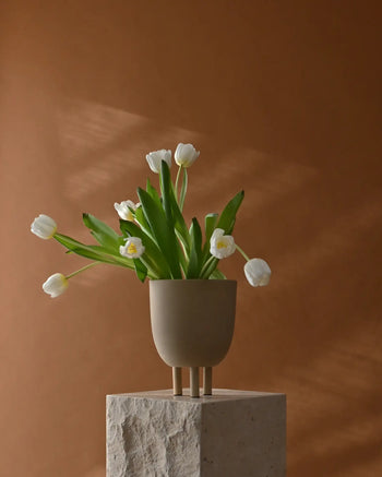 The 101 Copenhagen Duck Pot vase by 101 Copenhagen, featuring three short legs and a beige finish, displays white tulips with green leaves on a textured stone pedestal against a brown backdrop, bringing whimsical charm to minimalist décor.