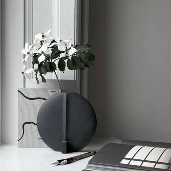 A minimalist scene features the 101 Copenhagen Guggenheim Vase, Petit—a round black, hand-glazed piece—holding white flowers with green leaves on a white surface, beside a pen, open book, and abstract art set against a gray and white background.