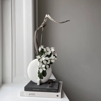 The 101 Copenhagen Guggenheim Vase, Petit by 101 Copenhagen, hand-glazed in white and styled with a dried leaf and white flowers, rests on two monochrome books by a window for a calm, modern Art Deco-inspired look.