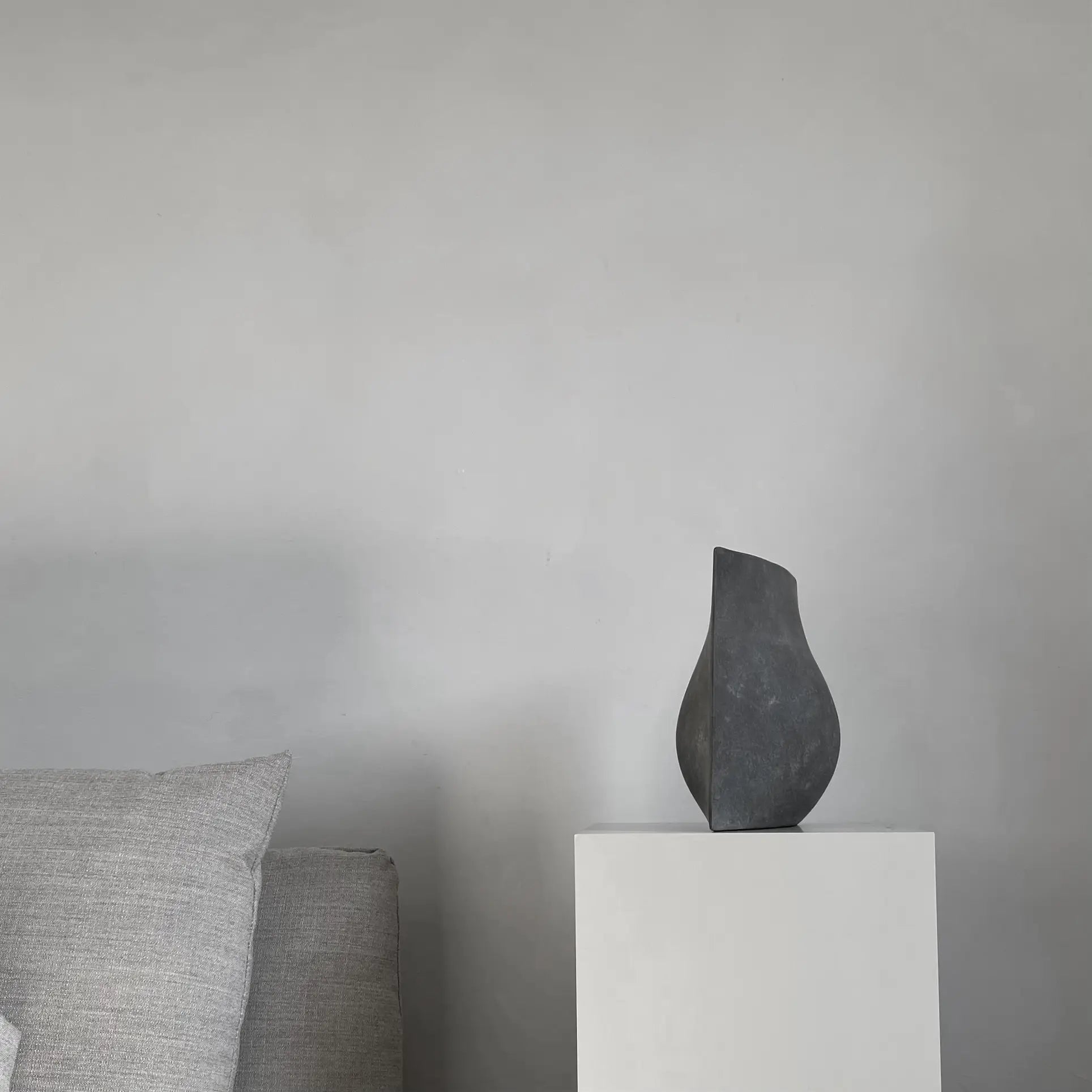 A minimalist scene with a light gray sofa and pillow, featuring the 101 Copenhagen Origami Vase, Mini by 101 Copenhagen on a white pedestal against a plain gray wall.