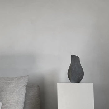 A minimalist scene with a light gray sofa and pillow, featuring the 101 Copenhagen Origami Vase, Mini by 101 Copenhagen on a white pedestal against a plain gray wall.