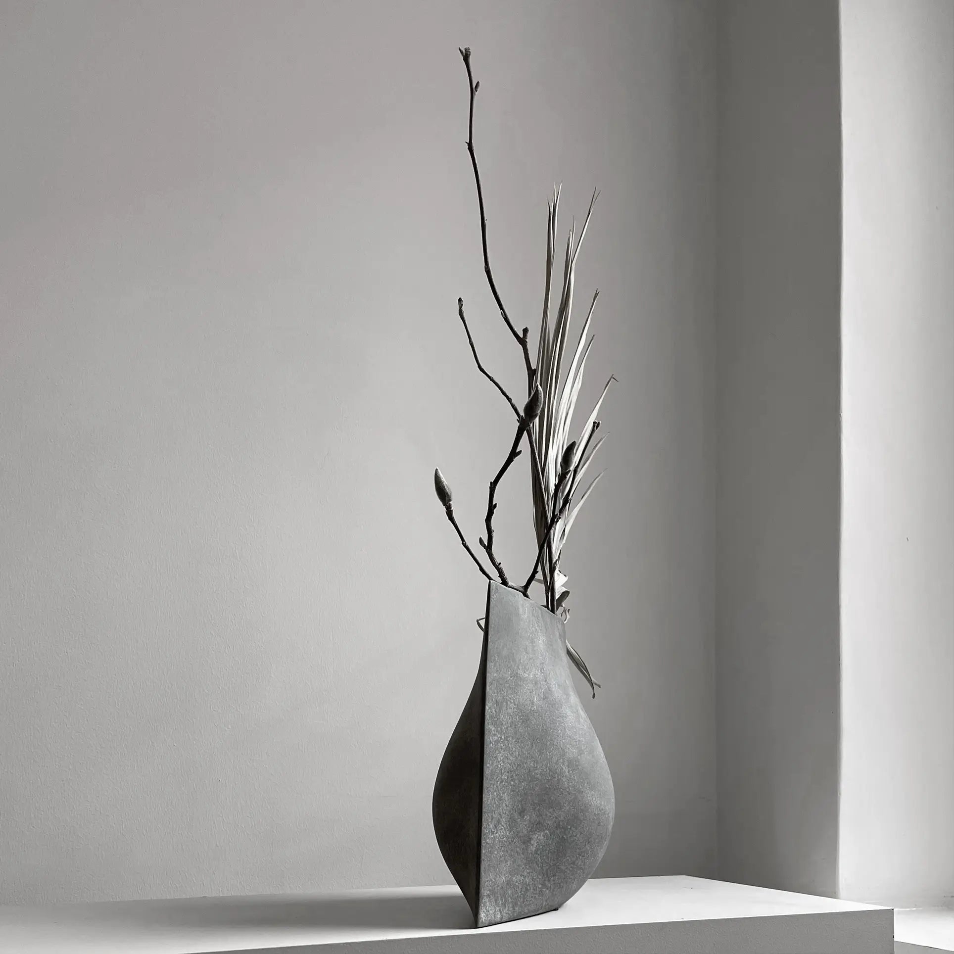 The 101 Copenhagen Origami Vase, Mini by 101 Copenhagen displays a few long, dried branches and grasses. This sculptural handmade vase sits on a pedestal in a softly lit room with smooth, neutral-toned walls.