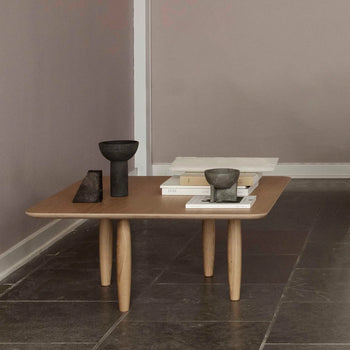 A minimalist wooden coffee table with rounded legs displays 101 Copenhagen Osaka Bowl, Mini, a stone sculpture, and stacked books on a tiled floor next to a mauve wall.