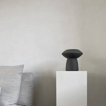A minimalistic scene with a light gray sofa on the left and the 101 Copenhagen Sphere Vase Fat by 101 Copenhagen as a striking centerpiece atop a white pedestal, lending timeless elegance to the plain, light-colored wall.