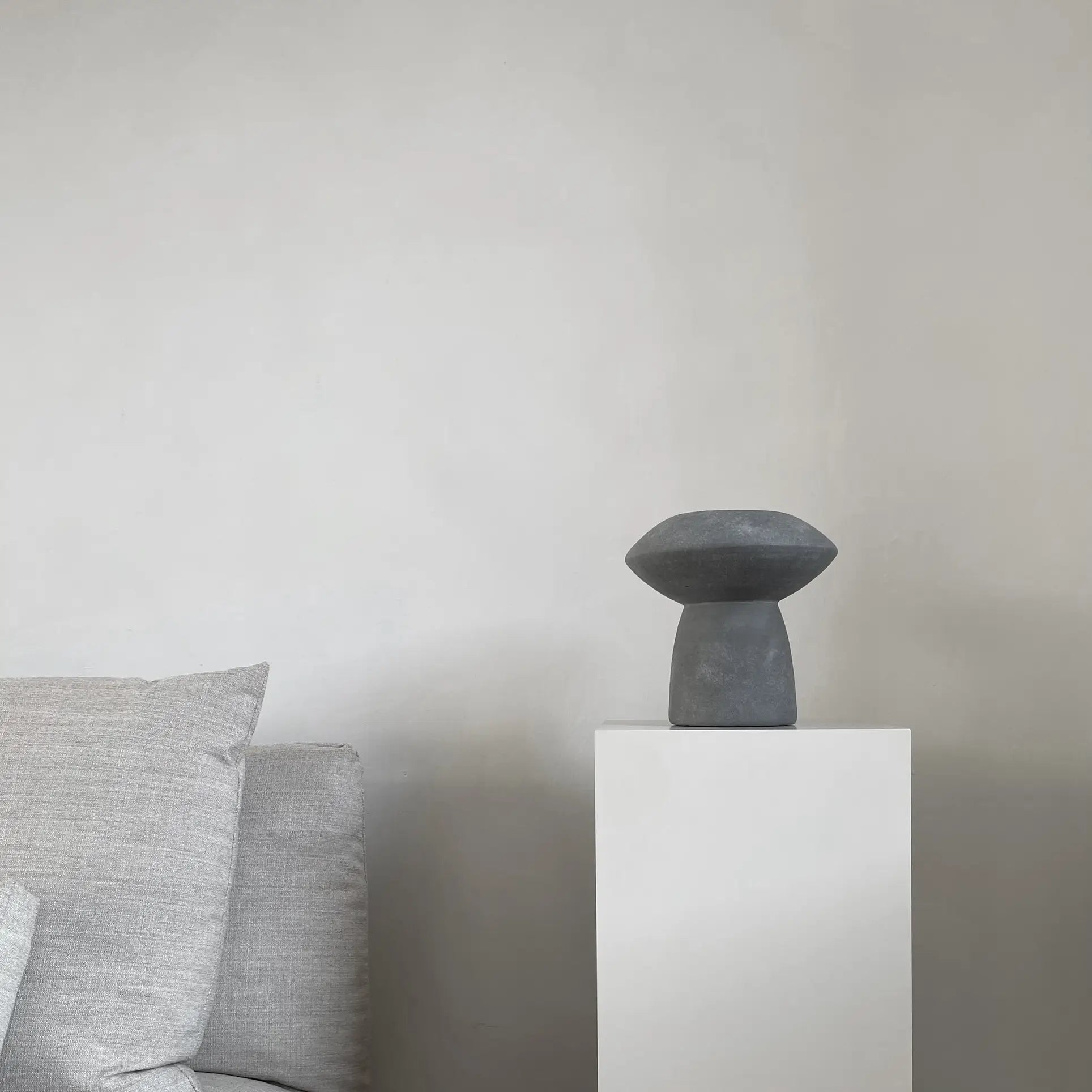 A minimalist room features a light gray sofa on the left and a white pedestal displaying the 101 Copenhagen Sphere Vase Fat, bringing timeless elegance from 101 Copenhagen against a plain, light-colored wall.