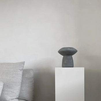 A minimalist room features a light gray sofa on the left and a white pedestal displaying the 101 Copenhagen Sphere Vase Fat, bringing timeless elegance from 101 Copenhagen against a plain, light-colored wall.