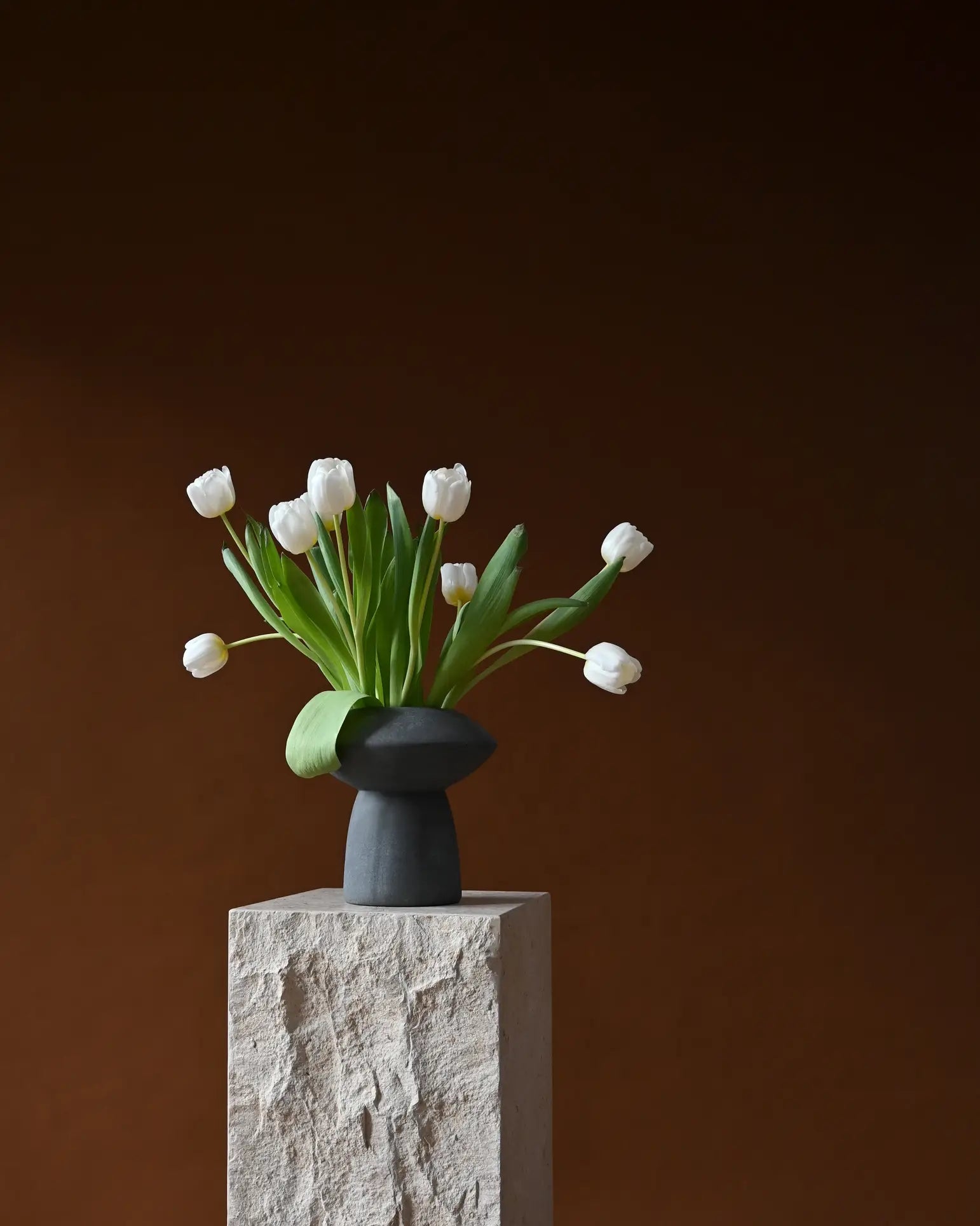 The 101 Copenhagen Sphere Vase Fat by 101 Copenhagen, filled with white tulips and green leaves, rests on a textured stone pedestal against a dark brown background, forming a captivating and timelessly elegant centerpiece.