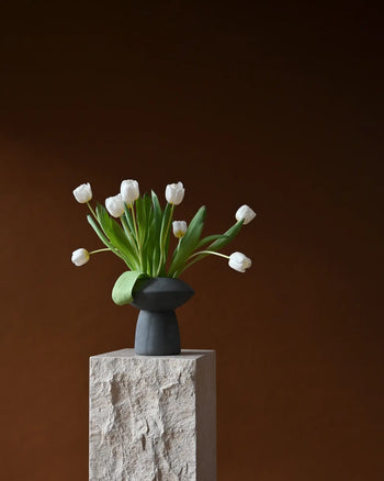 The 101 Copenhagen Sphere Vase Fat by 101 Copenhagen, filled with white tulips and green leaves, rests on a textured stone pedestal against a dark brown background, forming a captivating and timelessly elegant centerpiece.