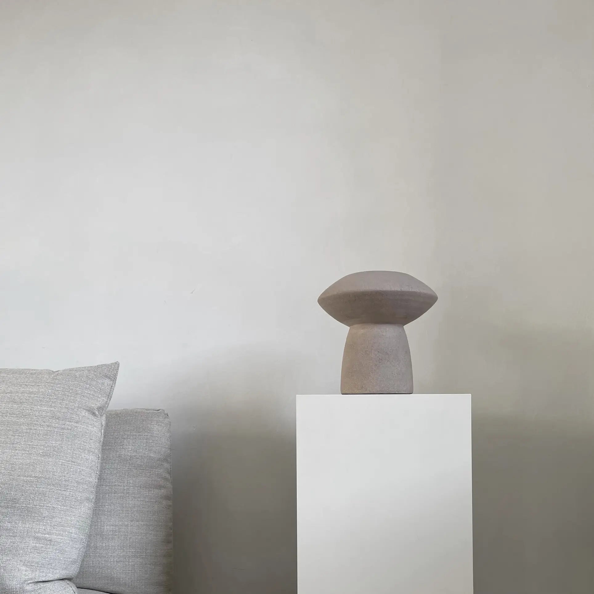 A minimalist scene with a light gray sofa, a cushion on the left, and the 101 Copenhagen Sphere Vase Fat by 101 Copenhagen as a striking centerpiece on a white pedestal, set against a plain, light wall.