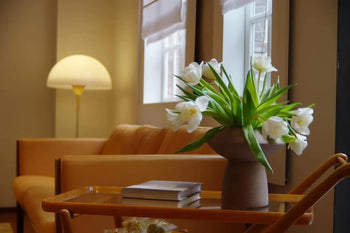 A cozy living room with a tan sofa, lit floor lamp, and wooden coffee table displaying books and the 101 Copenhagen Sphere Vase Fat by 101 Copenhagen—its white flowers creating a striking centerpiece near a window filled with natural light.