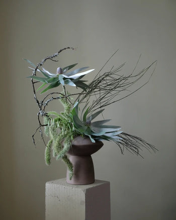 An eye-catching centerpiece, the 101 Copenhagen Sphere Vase Fat by 101 Copenhagen displays a minimalist floral arrangement with pale green leaves and branches, set on a textured pedestal against a muted background.