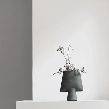 The 101 Copenhagen Sphere Vase Square, Mini—a minimalist black vase—holds white flowers and greenery on a white surface, set against white and gray walls for a sculptural look inspired by ancient Chinese vases.