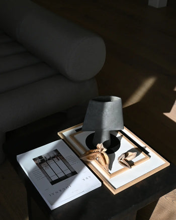 A black side table with sculptural elegance displays a 101 Copenhagen Sphere Vase Square, Mini with dried leaves, a gray lamp, framed art, and the book JAK JOURNAL beside a dark upholstered sofa in a sunlit room with wooden flooring.