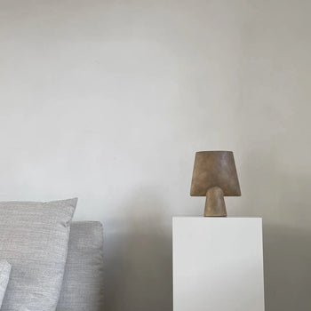 A minimalist scene with a gray couch and cushion on the left, and the 101 Copenhagen Sphere Vase Square, Mini displayed on a white pedestal—showcasing sculptural elegance from 101 Copenhagen against a plain light-colored wall.