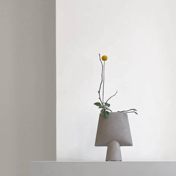 A minimalist scene showcases the 101 Copenhagen Sphere Vase Square, Mini in soft gray, holding a single branch with a yellow spherical flower and green leaves—an ideal modern décor accent against a plain white wall.