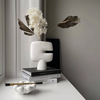 The 101 Copenhagen Tribal Vase, Mini by 101 Copenhagen, styled with dried flowers and palm leaves atop stacked books by a window, forms a minimal monochrome scene alongside a sculptural white ink brush holder.
