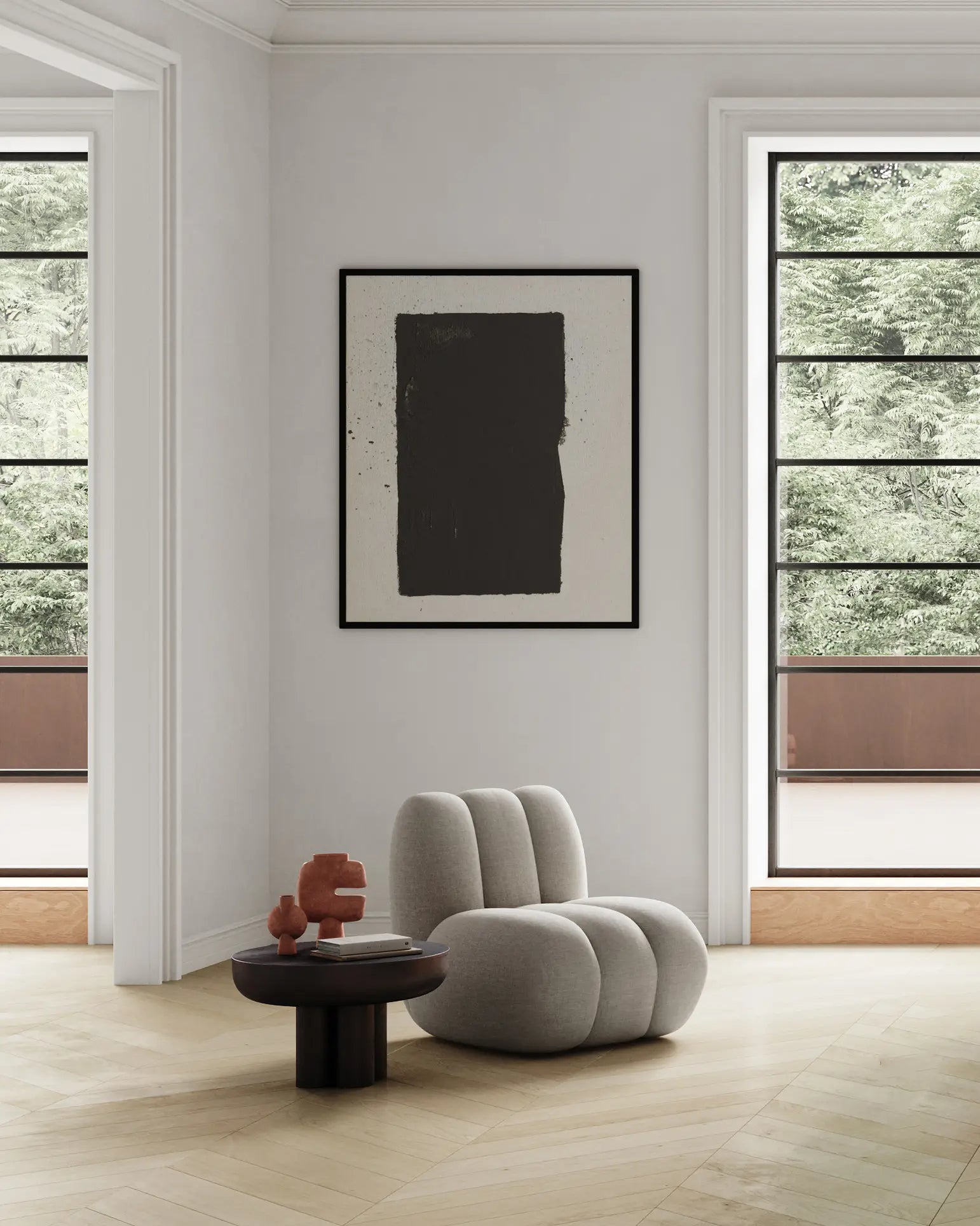 A minimalist living space with a plush, light grey chair and a round dark coffee table displaying the 101 Copenhagen Tribal Vase, Mini by 101 Copenhagen. A large abstract painting hangs on the wall while large windows overlook green trees.