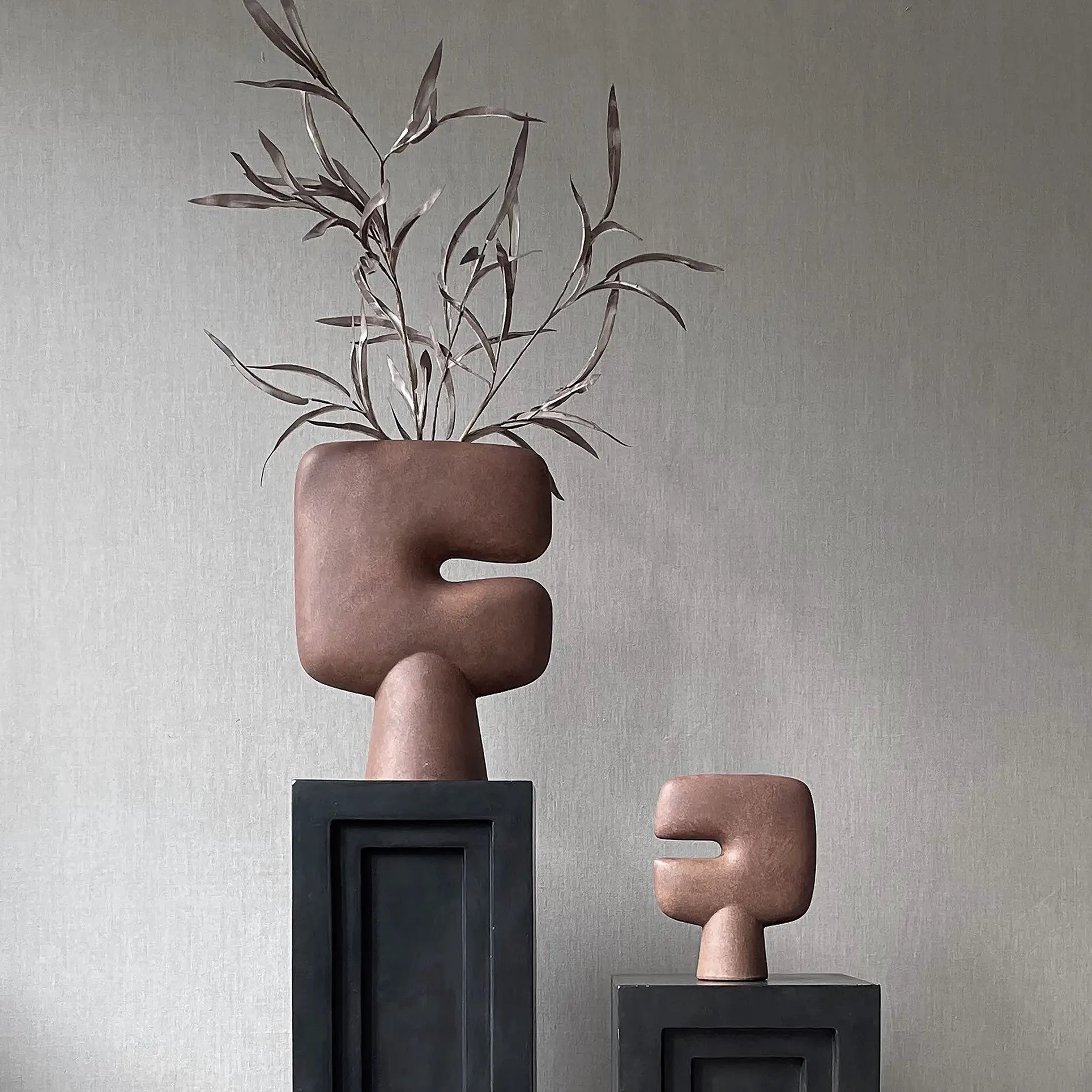 The 101 Copenhagen Tribal Vase, Mini by 101 Copenhagen, features an abstract brown sculptural form with geometric shapes. Displayed on a black pedestal beside a taller vase with branches, it sits against a neutral textured backdrop.