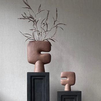 The 101 Copenhagen Tribal Vase, Mini by 101 Copenhagen, features an abstract brown sculptural form with geometric shapes. Displayed on a black pedestal beside a taller vase with branches, it sits against a neutral textured backdrop.