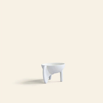 The 101 Copenhagen Wing Bowl, Mini by 101 Copenhagen is a small, minimalist white sculptural bowl with three uneven legs, crafted from fiber concrete and displayed on a light beige background.