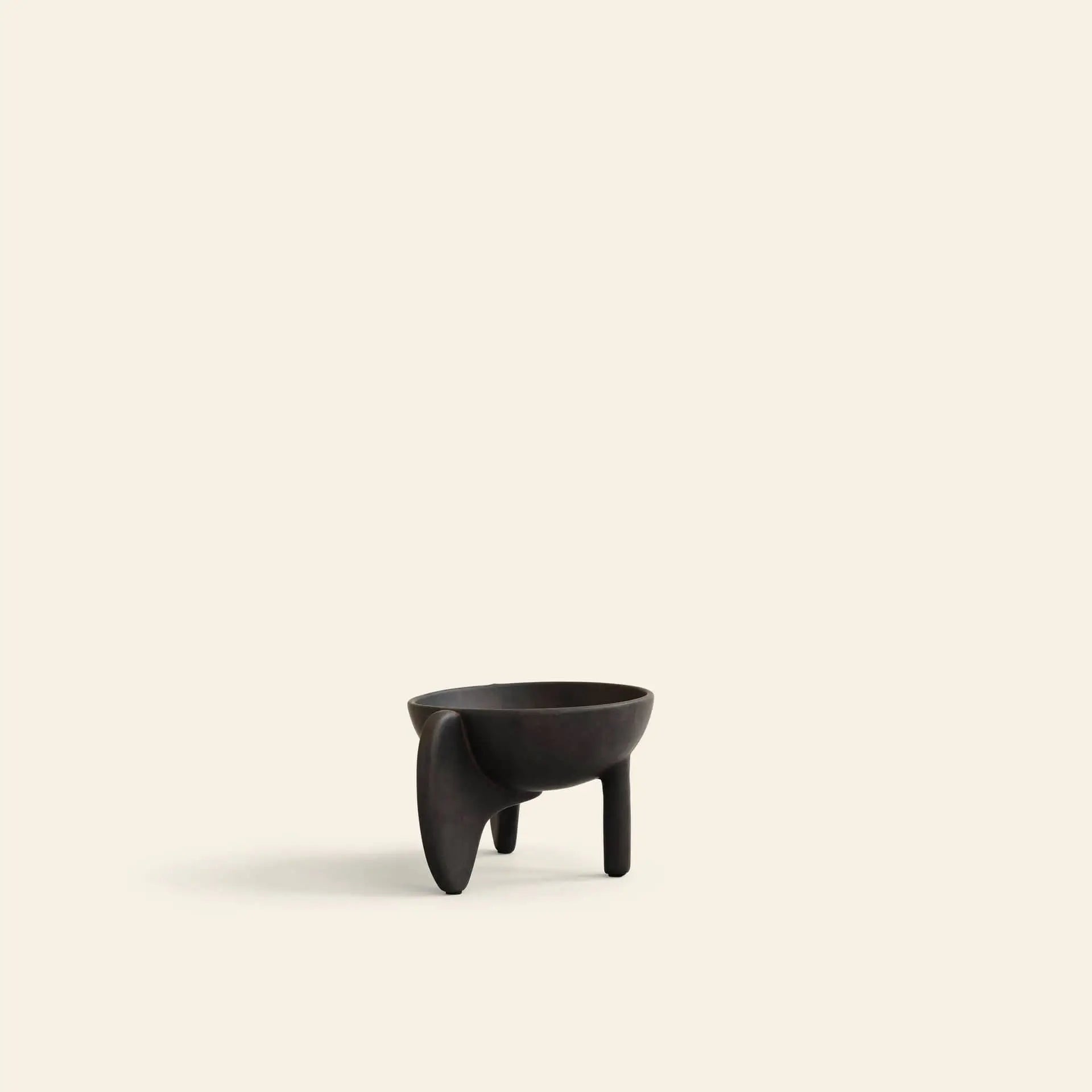 The 101 Copenhagen Wing Bowl, Mini by 101 Copenhagen is a small, dark fiber concrete bowl with an abstract sculptural design, featuring two simple legs and one larger curved leg. It sits on a plain light beige background.