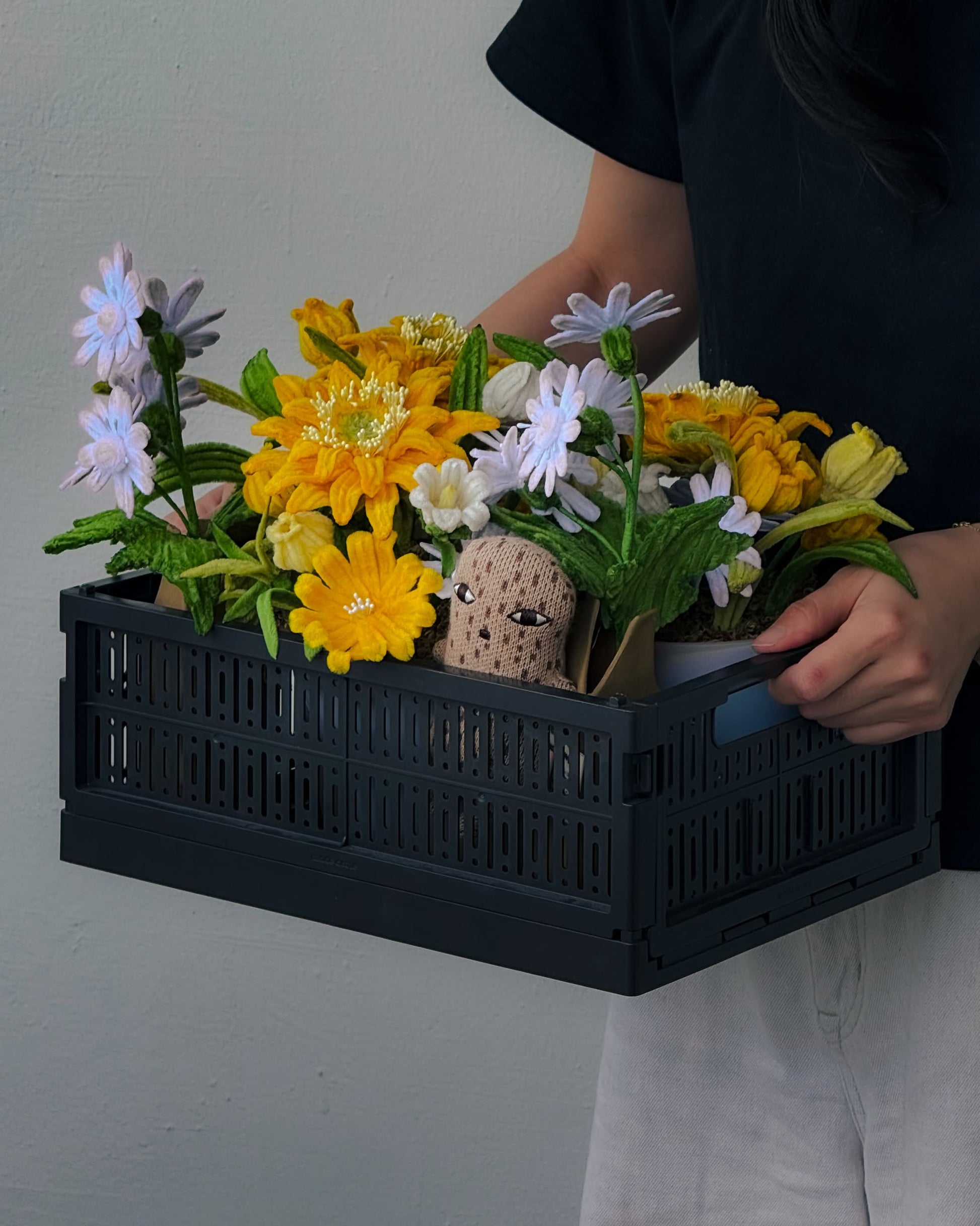 A person in a black shirt and white pants holds the Ah!GUGU x Stacked Store Bloom in Golden Hour - Handcrafted Flowers, featuring yellow and white artificial blooms and a plush toy, with florals standing out against a plain light-colored wall.
