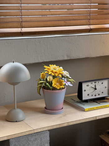 A small desk with a modern gray lamp, Ah!GUGU x Stacked Store Bloom in Golden Hour handcrafted flowers in a recycled pot with yellow and white blooms, a black and white clock, set against beige walls and window blinds.