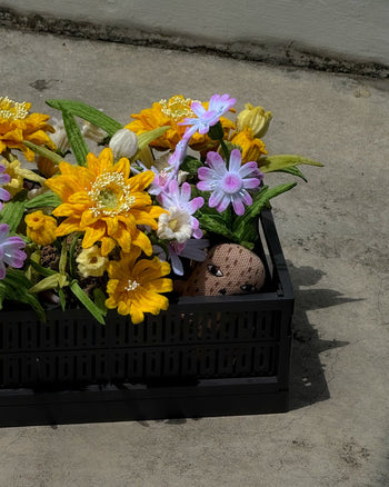 A black recycled plastic pot from Ah!GUGU holds the Ah!GUGU x Stacked Store Bloom in Golden Hour – Handcrafted Flowers, featuring artificial yellow, white, and light purple blooms. A painted rock with a face peeks out among the florals.