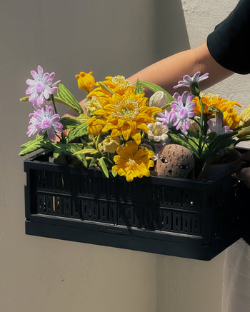 A person holds the Ah!GUGU x Stacked Store Bloom in Golden Hour - Handcrafted Flowers by Ah!GUGU, featuring colorful crocheted flowers and a small crocheted figure in a black crate, set against a light background.