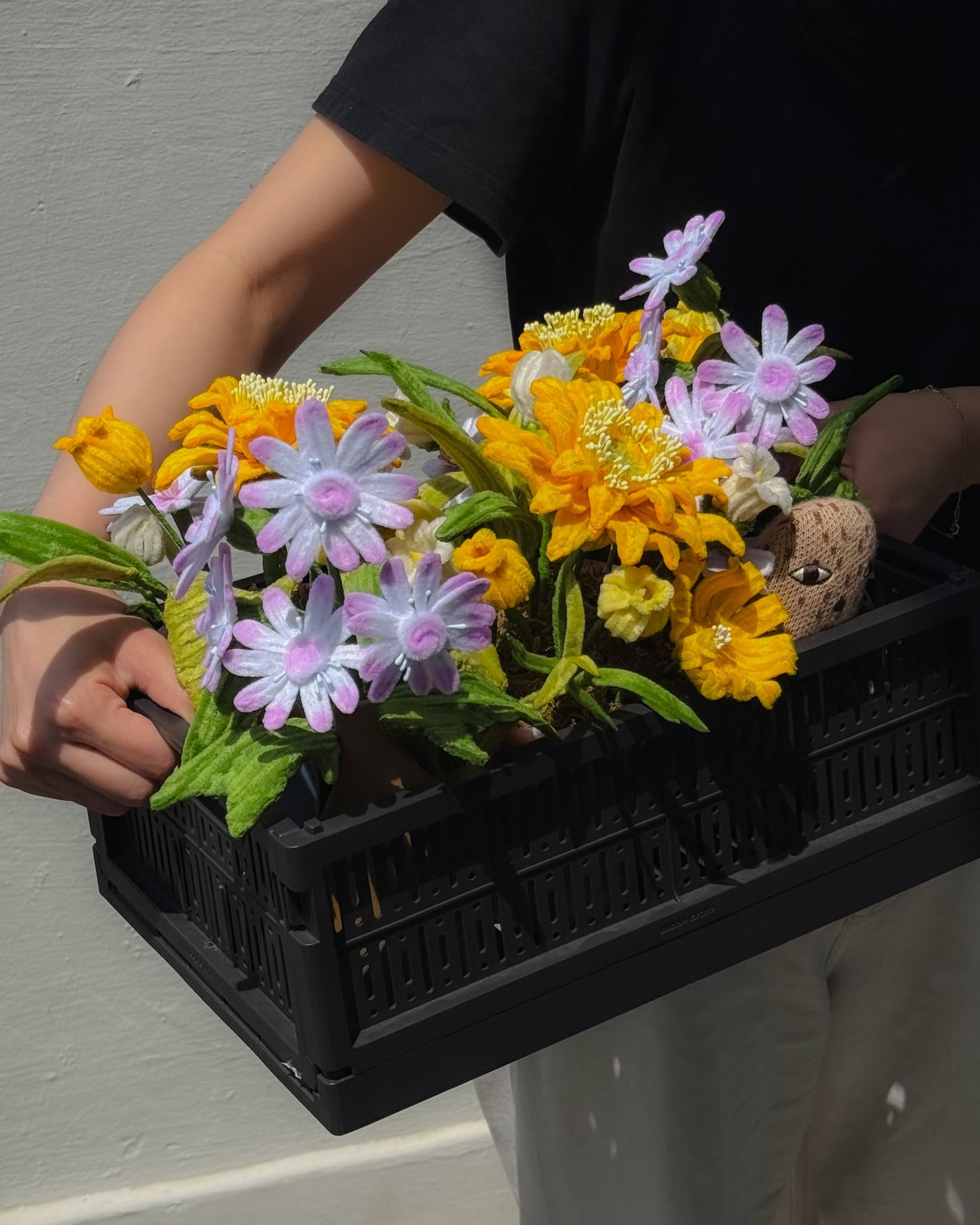A person in a black shirt holds the Ah!GUGU x Stacked Store Bloom in Golden Hour—Handcrafted Flowers, a black crate filled with florals and greenery by Ah!GUGU, while standing against a light-colored wall.