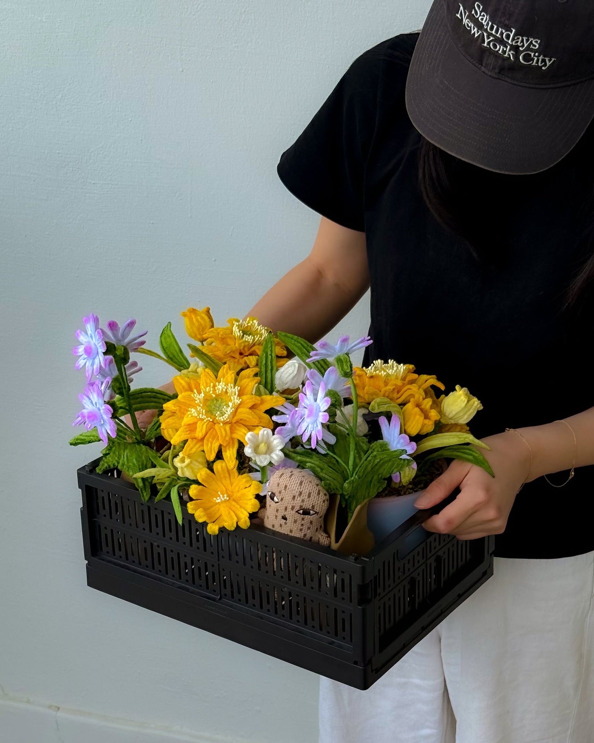A person wearing a black cap, black shirt, and white pants holds an Ah!GUGU x Stacked Store Bloom in Golden Hour - Handcrafted Flowers arrangement against a plain light-colored wall.