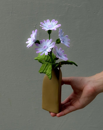 A hand holds the Ah!GUGU x Stacked Store Still Life in Bloom - Handcrafted Flowers, featuring delicate light purple fabric flowers with green leaves in a sculptural vase, set against a plain gray background.