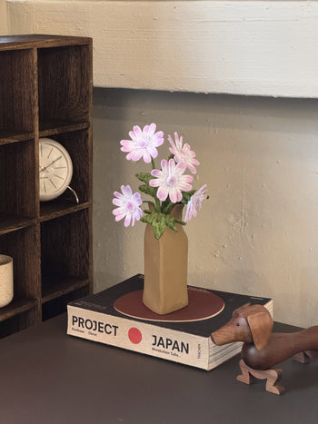 The Ah!GUGU x Stacked Store Still Life in Bloom - Handcrafted Flowers are displayed in a sculptural vase on a book, with a wooden dog figurine and a shelf holding a small clock in the background.