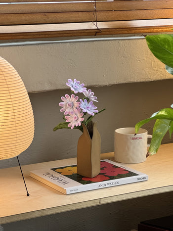 An Ah!GUGU x Stacked Store Still Life in Bloom - Handcrafted Flowers vase sits on a wooden shelf beside an “I LOVE YOU” mug and Andy Warhol book, near a paper lantern, green plant, and under wooden blinds.