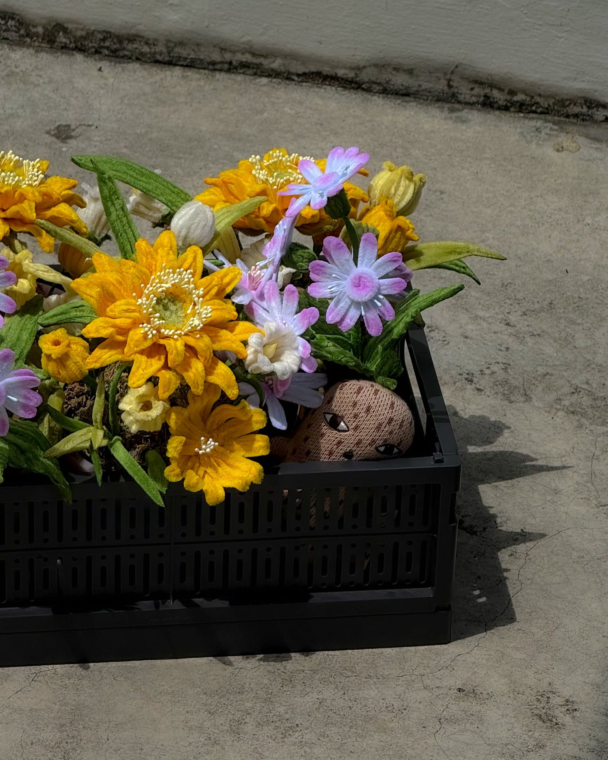 The Ah!GUGU x Stacked Store Still Life in Bloom - Handcrafted Flowers features a black crate vase with vibrant artificial blooms, accented by a cute face-painted rock, creating a playful arrangement perfect for any surface.